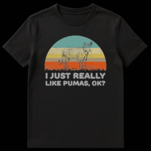 Vintage-style t-shirt graphic featuring a stylized puma with blue eyes standing on grass, set against a distressed multi-color sunset. Below the graphic, text reads "I JUST REALLY LIKE PUMAS, OK?" in a distressed grey font on a black background. on Black t-shirt